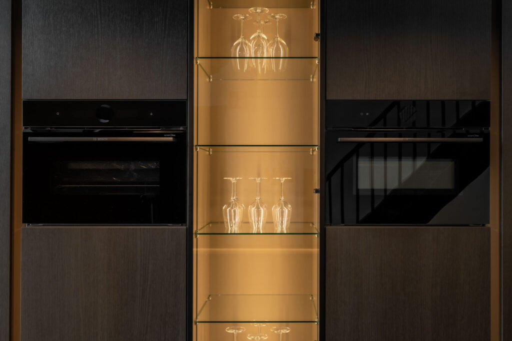 Hoge kastenwand met inbouwovens en een verlichte glazen vitrine voor wijnglazen